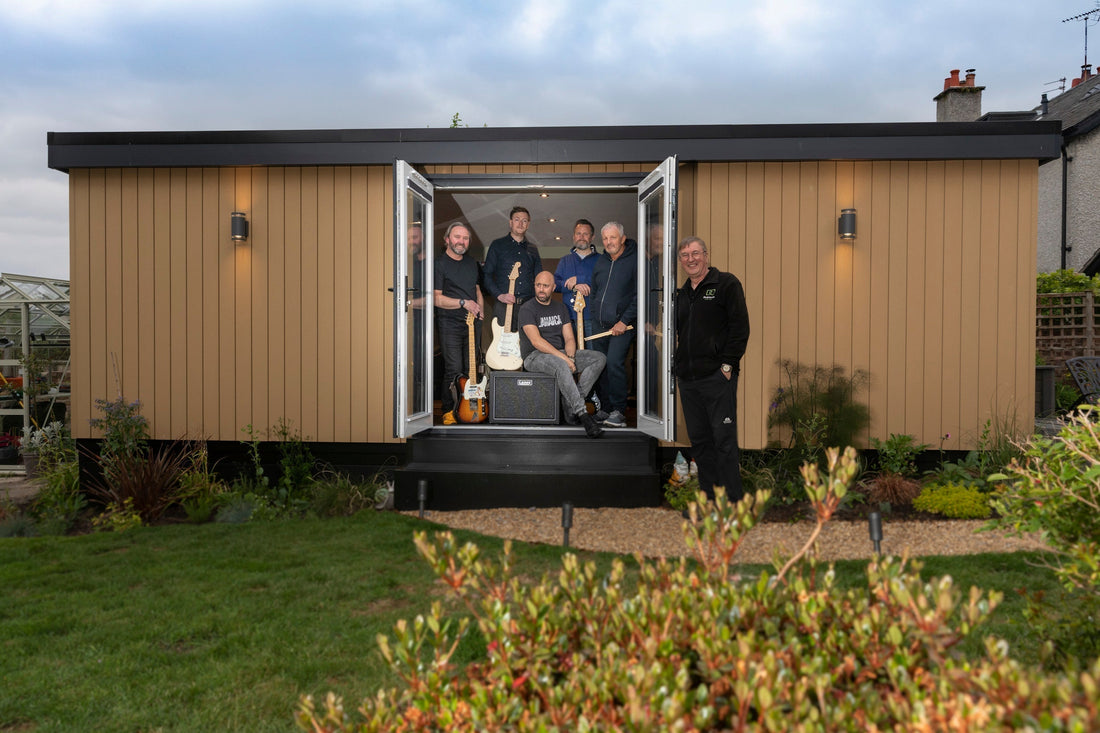 A garden room with brown composite cladding, two external lights, in a garden, with the band and rubicon director John posing for a picture in the doorway.