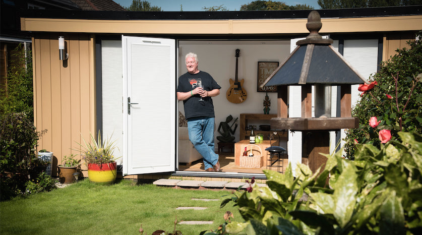 A man standing in front of a small shed in a garden.