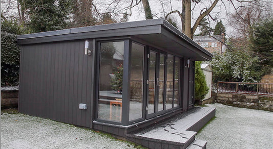 A Rubicon Garden Rooms Vista Style garden shed with a glass door, snow on the ground, and a self-coloured render.