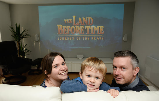A family sits on a couch in front of a projector showing the land before time, in their Rubicon Garden Room, used as a Home Cinema.