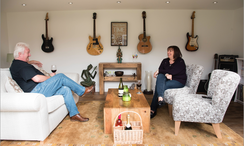 A man and woman sitting in a living room with guitars on the wall.